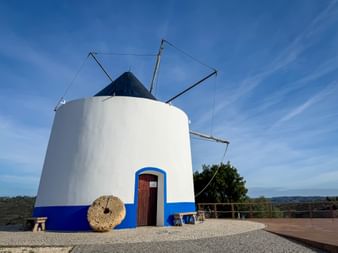 Weiße Windmühle mit blauem Rand und dunklem Kegeldach in Odeceixe, Alentejo. Die Struktur hat Holzflügel und einen Mühlstein neben dem Eingang.
