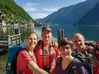 Group of five hikers with backpacks posing at Hallstatt lakeside. Historic village and mountains visible in background under blue sky.