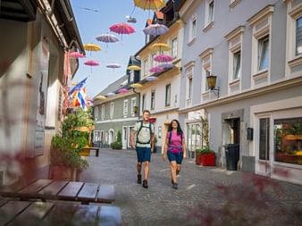 Two hikers with backpacks walking through Villach's pedestrian zone decorated with colorful umbrellas hanging overhead between historic buildings.