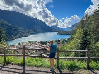 Female hiker with blue backpack at wooden fence viewpoint overlooking Lake Molveno, village, and mountains under dramatic cloudy sky.