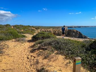 Hiker with backpack on sandy trail overlooking cliffs and blue ocean on Rota Vicentina. Trail marker visible in foreground under clear sky.