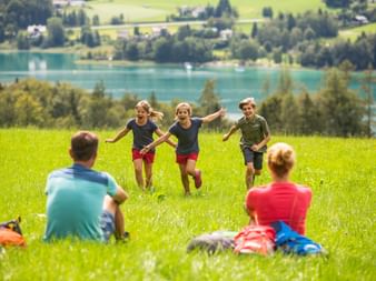 Family on green meadow in Salzkammergut: three children running toward two seated adults, with turquoise lake and mountains in background.