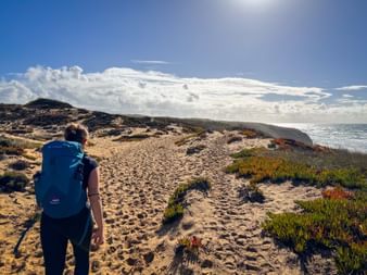 Wanderin mit blauem Rucksack auf sandigem Küstenpfad am Praia do Brejo Largo an der Rota Vicentina, mit Meerblick und dramatischen Wolken.