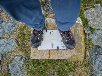Füße eines Wanderers in blauer Hose und Wanderschuhen auf einem weißen Grenzstein, eingebettet in felsigen Boden mit Gras und Moos.