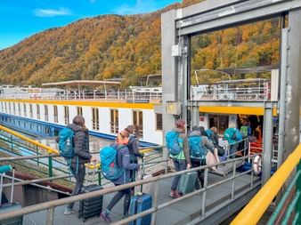 Group of tourists with backpacks boarding a white and orange river cruise ship on the Danube. Autumn hills with colorful foliage visible in background.