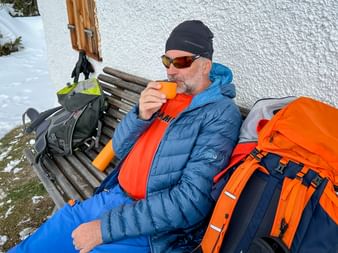 Wanderer in blauer Jacke und Sonnenbrille trinkt aus oranger Tasse auf Holzbank. Oranger Rucksack neben ihm, schneebedeckter Boden und weiße Wand dahinter.