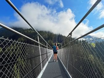 Wanderer mit Rucksack überquert eine Hängebrücke am Lechweg, umgeben von bewaldeten Bergen und blauem Himmel mit Wolken.