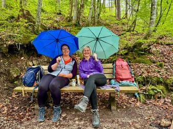 Two female hikers sitting on a wooden bench in a lush forest, holding blue and turquoise umbrellas. Backpacks rest beside them on the bench.