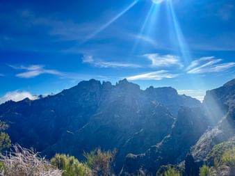 Dramatic mountain panorama of Pico Ruivo in Madeira with jagged peaks under bright blue sky with sunbeams and wispy clouds.