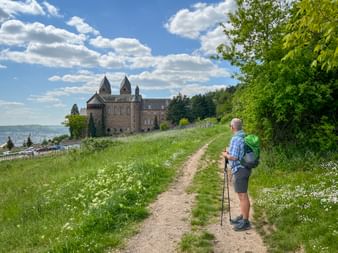 Hiker with backpack on path approaching St. Hildegard Abbey in Rüdesheim. Stone monastery with towers visible on hillside above the Rhine.