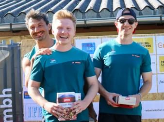 Three smiling men in turquoise sports shirts holding prize boxes at Grabenseelauf 2025. Ray, Manuel and Nico pose together outdoors.