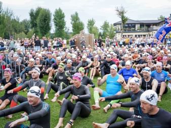 Large group of triathletes in wetsuits and swim caps sitting on grass before the Trumer Triathlon start, with spectators and buildings in background.