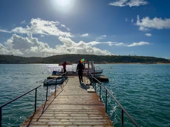 Holzsteg mit Metallgeländer über türkisfarbenem Wasser in Vila Nova de Milfontes. Zwei Personen gehen auf ein Fährboot zu unter blauem Himmel.