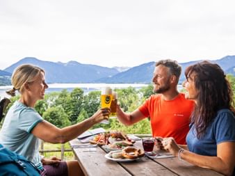Three hikers toasting with beer at a wooden table with Bavarian snacks, overlooking a lake and mountains on the König Ludwig Weg trail.