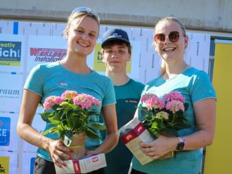 Three smiling women in turquoise sports shirts holding flower bouquets and awards at Grabenseelauf 2026, standing before sponsor backdrop.