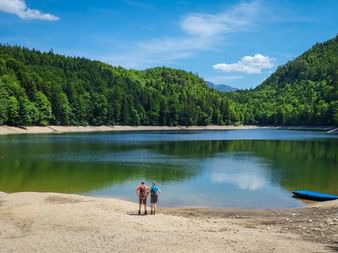 Two hikers with backpacks standing on sandy shore of Nussensee, surrounded by forested hills under blue sky with white clouds.