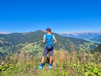 Wanderin mit blauem Rucksack auf der Schmittenhöhe mit Blick auf grüne Alpentäler und schneebedeckte Berge bei Zell am See im Pinzgau.