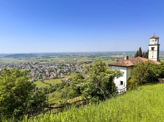 Weiße Kirche mit Glockenturm am Hang mit Blick über das Friaul-Tal mit Stadt unten, umgeben von grünen Feldern und Weinbergen unter blauem Himmel.