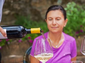 Weißwein wird aus einer Flasche in ein Glas in der Toskana eingeschenkt, im Hintergrund eine Frau in pinkem Shirt zwischen Grünpflanzen.