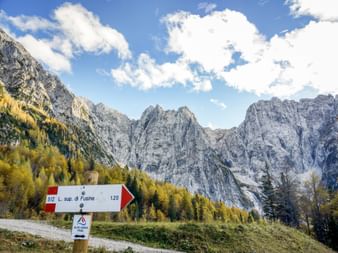 Hiking trail sign pointing to Laghi di Fusine with dramatic rocky mountain peaks and autumn forest in the background under blue sky with clouds.