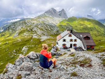 Female hiker sitting on rocks overlooking Leutkircher Hütte, a mountain hut with colorful shutters, surrounded by green Alpine meadows and peaks.