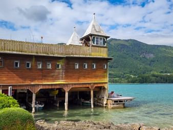 Wooden boathouse with tower on Fuschlsee with turquoise water, boats underneath, and green mountains in the background under cloudy sky.