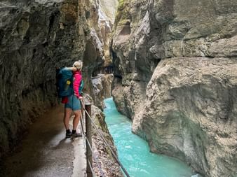 Wanderin mit Rucksack auf Steg in der Höllentalklamm, blickt auf türkisfarbenen Gletscherbach in enger Felsschlucht.