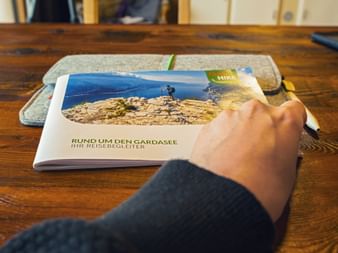 Hand reaching for Lake Garda travel guide on wooden table. The brochure shows a hiker on rocky terrain with blue lake view. A gray felt folder lies behind.