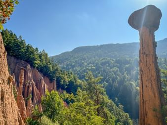 Tall earth pyramids with stone caps at Ritten, South Tyrol. Reddish clay pillars rise against forested mountains under blue sky.