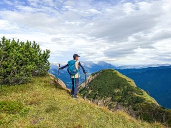 Wanderin mit blauem Rucksack und Wanderstöcken auf grasbewachsenem Bergrücken im Pinzgau, Österreich, mit Alpengipfeln im Hintergrund.
