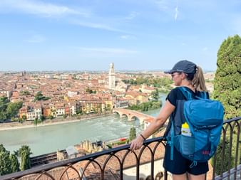 Female hiker with blue backpack viewing Verona's old town from viewpoint. The Adige River and historic bridge are visible below the terracotta rooftops.