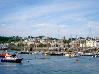 Hafenansicht von St Peter Port mit Segelbooten und Lotsenboot im Vordergrund, bunte Gebäude am Hang mit sichtbarer Kirchenkuppel.