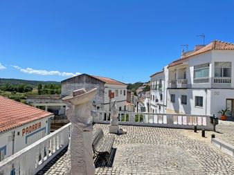 Kopfsteinpflasterplatz in Aljezur mit weißer Balustrade, traditionellen weißen Gebäuden mit Terrakottadächern und Hügeln im Hintergrund.