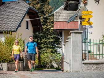 Two hikers walking past yellow trail signs in Kranjska Gora village with alpine houses and mountains in the background.