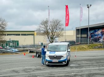 White Renault van on training course with orange cones. Person in blue jacket stands beside vehicle on marked parking lot. Eurofun flags visible.
