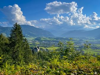 Panoramic view of Saalfelden valley with green meadows and town, framed by mountains and dramatic white clouds in blue sky.