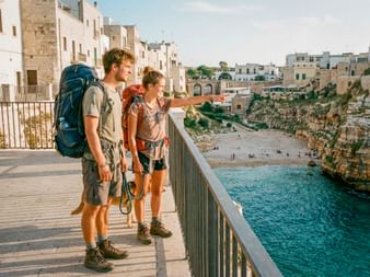 Zwei Wanderer mit Rucksäcken auf einer Promenade in Polignano a Mare, Apulien, mit Blick auf einen kleinen Strand zwischen weißen Gebäuden und Felsen.