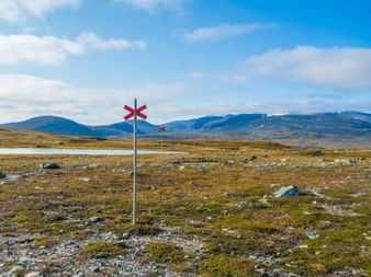 Rote Wegmarkierungen auf Pfählen in Funäsfjällen-Tundralandschaft mit Moos, Felsen und fernen Bergen unter blauem Himmel in Jämtland.
