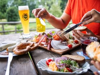 Person im orangenen Shirt genießt traditionelle bayerische Brotzeit im Freien mit Bierglas, Holzbrett mit Aufschnitt, Käse und Brot.