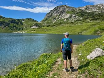 Wanderer mit blauem Rucksack steht am Formarinsee und blickt auf klaren Bergsee, umgeben von grünen Almwiesen und felsigen Gipfeln.