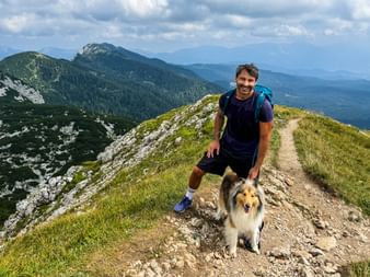 Lächelnder Wanderer mit Rucksack und Collie-Hund auf Bergpfad am Visevnik in den Julischen Alpen. Bewaldete Gipfel und Täler im Hintergrund.