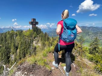 Wanderin mit blauem Rucksack blickt auf das Jakobskreuz in den Kitzbüheler Alpen mit Bergpanorama und Wäldern unter blauem Himmel.