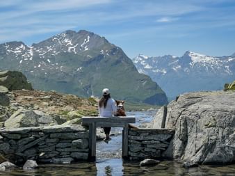 Frau mit Hund auf Steinbrücke über Bergbach am Rusettasee auf der Via Spluga, mit schneebedeckten Alpengipfeln im Hintergrund.