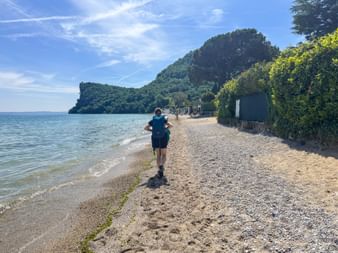 Wanderer mit Rucksack auf Schotterweg am Gardasee-Ufer bei Salò, mit bewaldeten Bergen und blauem Himmel im Hintergrund.