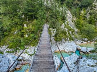 Hölzerne Hängebrücke über die türkisfarbene Soča in Trenta, Julische Alpen. Felsschlucht mit üppiger grüner Vegetation auf beiden Seiten.