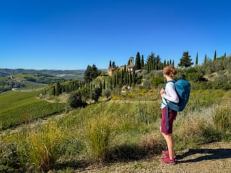Woman with backpack viewing Tuscan hills with cypress trees and a villa. Rolling green fields and blue sky in the background.
