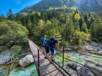 Drei Wanderer mit Rucksäcken überqueren eine Holzbrücke über türkisfarbenen Fluss in Lepena, Julische Alpen. Dichter Wald und Berge im Hintergrund.