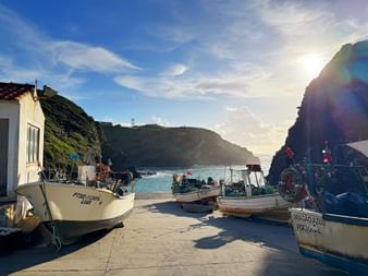 Fischerboote auf Betonpier im Hafen Porto das Barcas im Alentejo. Felsige Klippen rahmen eine türkisfarbene Bucht unter blauem Himmel.