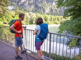 Zwei Wanderer mit Rucksäcken stehen an einem Geländer mit Blick auf den Lechfall und Fluss, umgeben von bewaldeten Bergen.