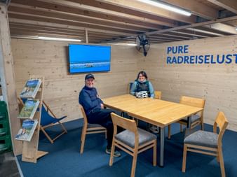 Two cyclists sitting at wooden table in modern visitor center with exposed ceiling beams, blue wall screen, and 'PURE RADLEISELUST' signage.
